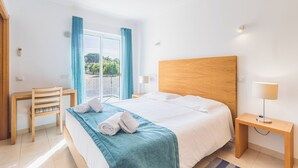 Quarto de hotel com cama, escrivaninha, cadeira, mesa de cabeceira e janela com cortinas.
