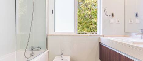 Un baño moderno con ducha de vidrio, un bidé, un lavabo y una ventana con vista a la vegetación.