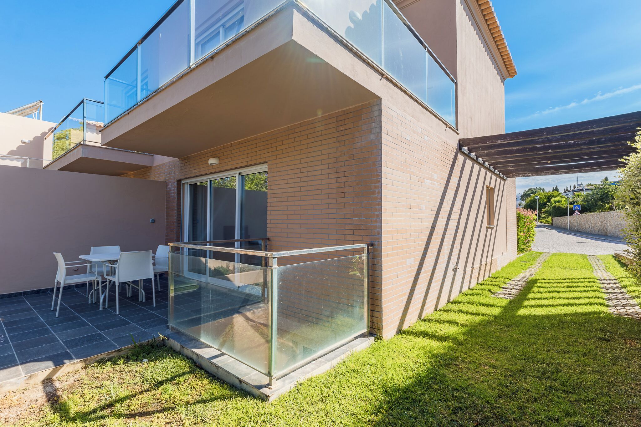 Una casa moderna con un balcón de vidrio, un patio con sillas y un césped bien cuidado.