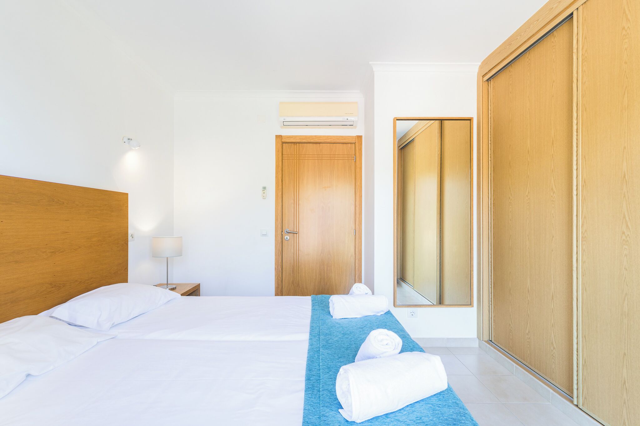 Hotellihuone, jossa on puinen sängynpääty, valkoisilla lakanoiden peitetty sänky, sininen pyyhe lattialla, puinen vaatekaappi ja peilillä varustettu ovi.