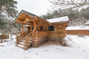 Uma cabana de tronco com escada de madeira e varanda, cercada por terreno e árvores cobertos de neve.