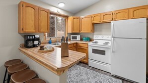 A kitchen with wooden cabinets, a white refrigerator, a stove, a microwave, and a coffee maker. There are stools and a rug on the floor.