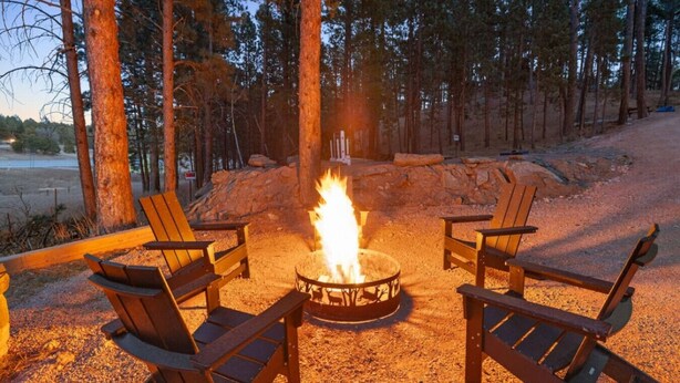 A campfire with four Adirondack chairs arranged around it in a forested area.