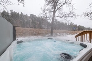 A hot tub with a wooden deck and a view of a snowy landscape with trees.