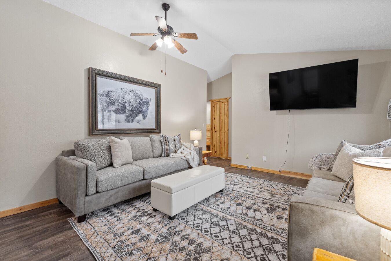 A living room with a gray sofa, a white ottoman, a patterned rug, a ceiling fan, a framed picture, and a flat-screen TV.