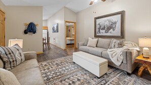 A spacious living room with a sofa, a coffee table, and a patterned rug. There is a ceiling fan, a framed picture on the wall, and a door leading to another room.