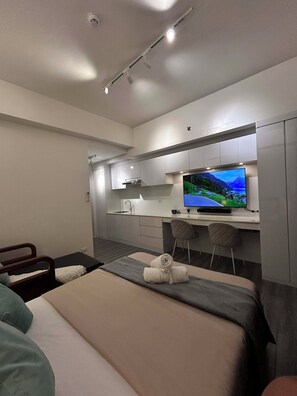 A modern hotel room with a bed, a desk with a flat-screen TV, a small kitchenette, and a bathroom.