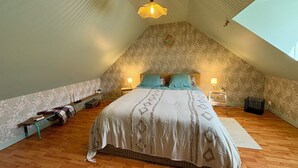 Quarto com teto inclinado, cama com roupa de cama estampada, duas luminárias de cabeceira, um banquinho com livros e uma mesinha com abajur.