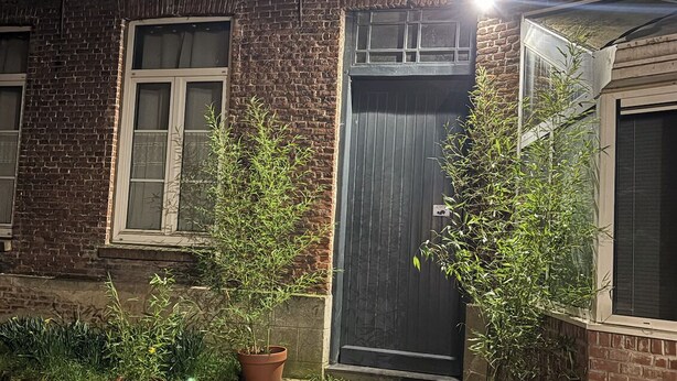 Edifício de tijolos com porta de entrada escura, plantas em vasos e um caminho de pedras que leva à porta.