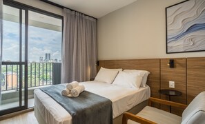 A hotel room with a large window, a bed with white linens, a grey throw blanket, and a city view. - Studio in Condo w/ Gym in Consolacao Mac0046 (São Paulo)