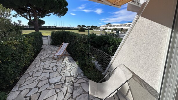 A stone-paved patio with a striped lounge chair, a pergola overhead, and a view of a landscaped area with a tree and distant buildings. - RESIDENCE L'ISLE CATHERINE BEL APPARTEMENT 2 PIECES RDC JARDIN PRIVATIF PK ET CLIM (Le Grau-du-Roi)