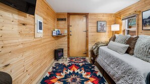 Une pièce chaleureuse aux murs en bois, avec un lit recouvert d’une literie à motifs, un tapis coloré, une petite table et un téléviseur fixé au mur.