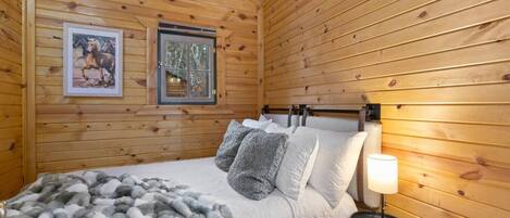 A wooden cabin room with a bed, pillows, a blanket, a framed picture, and a nightstand with a lamp.