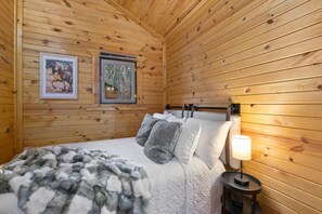 Puinen mökkimäinen huone, jossa on sänky, tyynyjä, peitto, kehystetty kuva ja yöpöytä, jossa on lamppu.