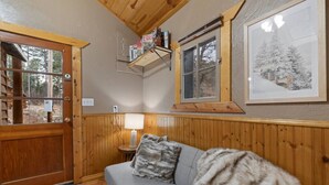 A cozy room with a wooden door, a grey sofa with a fur throw, a lamp, and a framed picture of a snowy landscape. - Angler’s Haven Cabin: Black Hills Quiet Escape (Rapid City)