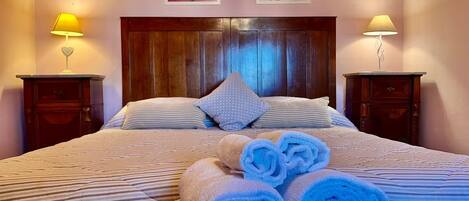 A bedroom with a wooden headboard, two bedside tables with lamps, a bed with striped bedding, and rolled towels on the floor.