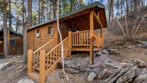 Ein rustikales Blockhaus mit einer Holztreppe, die zu einer Veranda führt, umgeben von Bäumen und felsigem Gelände.