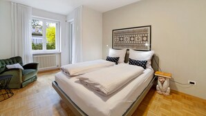 Une chambre à coucher avec un lit, un fauteuil vert, une fenêtre avec des rideaux et une œuvre d’art murale.