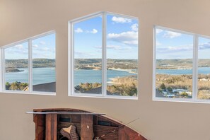 A room with three large windows offering views of a lake, a bridge, and surrounding landscape. - Luxury Lakeview Cabin + Lazy River & Pickleball (Kimberling City)