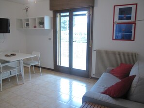 Un salón con sofá blanco, una mesa de comedor con sillas, televisor en la pared y puerta corrediza de vidrio que da a un área exterior.