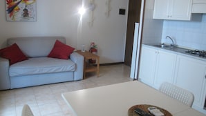 A living room with a grey sofa, red cushions, a small wooden table, and a kitchen area with white cabinets and a sink.