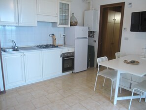 Una cocina compacta con armarios blancos, estufa a gas, horno integrado, refrigerador y comedor con mesa y sillas.