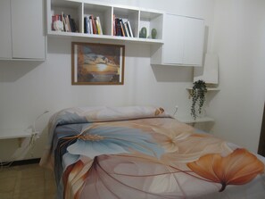 Un dormitorio con una cama, una estantería, un cuadro enmarco y una planta pequeña.