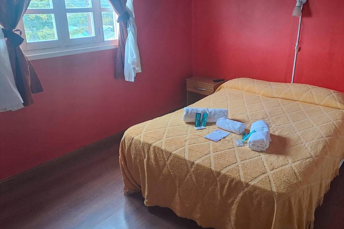 Een eenpersoonsbed met een gele sprei, twee opgerolde handdoeken en een klein nachtkastje met een lamp.