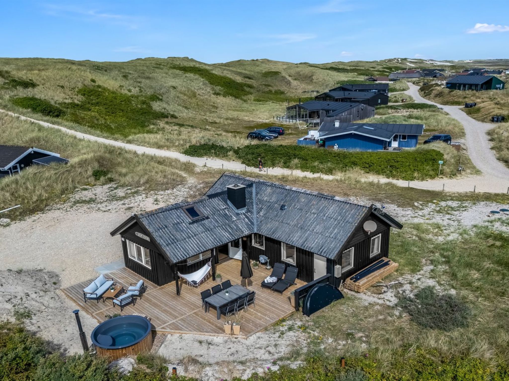 A wooden house with a deck and outdoor seating, surrounded by dunes and other buildings.