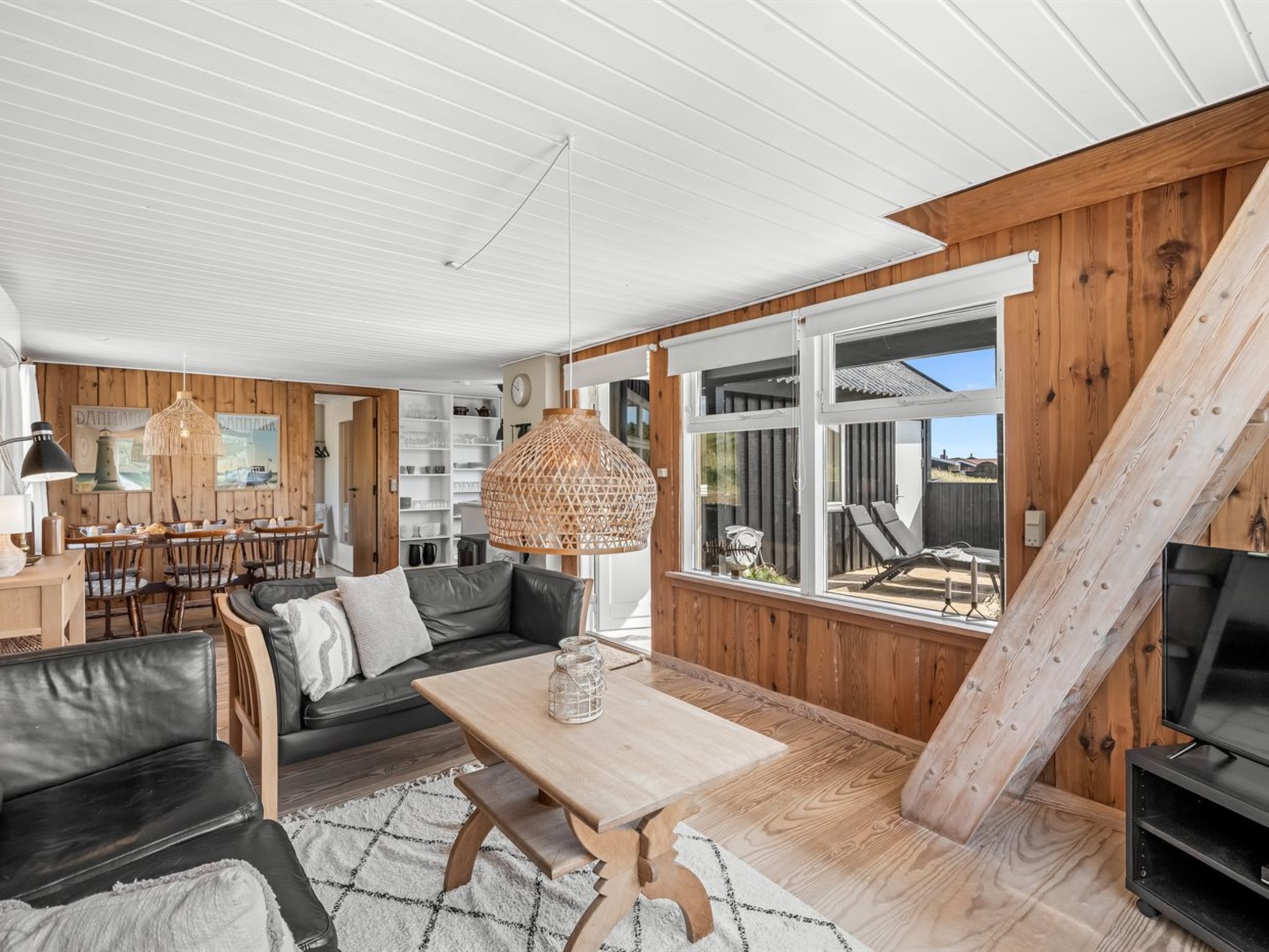 A modern living room with a wooden interior, a dining area, and a large window offering a view of the outdoors.