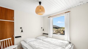 A bedroom with a bed, a chair, a window with curtains, and a hanging light fixture. - "Monna" -  from the sea by Interhome (Hvide Sande)
