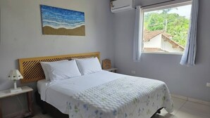 Ein Schlafzimmer mit Bett, Nachttisch, Lampe, Gemälde, Klimaanlage und einem Fenster mit Vorhängen.
