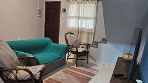 Ein kleiner, gut beleuchteter Raum mit einem grünen Sofa, einem Korbstuhl, einem Holzschrank und einem Fernseher.