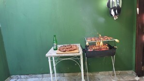 Ein kleiner Grillbereich im Freien mit Tisch, auf dem sich eine Bierflasche und ein Teller mit Essen befinden, vor einer grünen Wand.