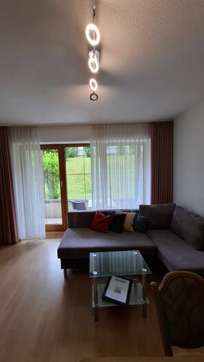 Ein Wohnzimmer mit einer Couch, einem Glastisch und Blick auf einen Garten durch eine Schiebetür.
