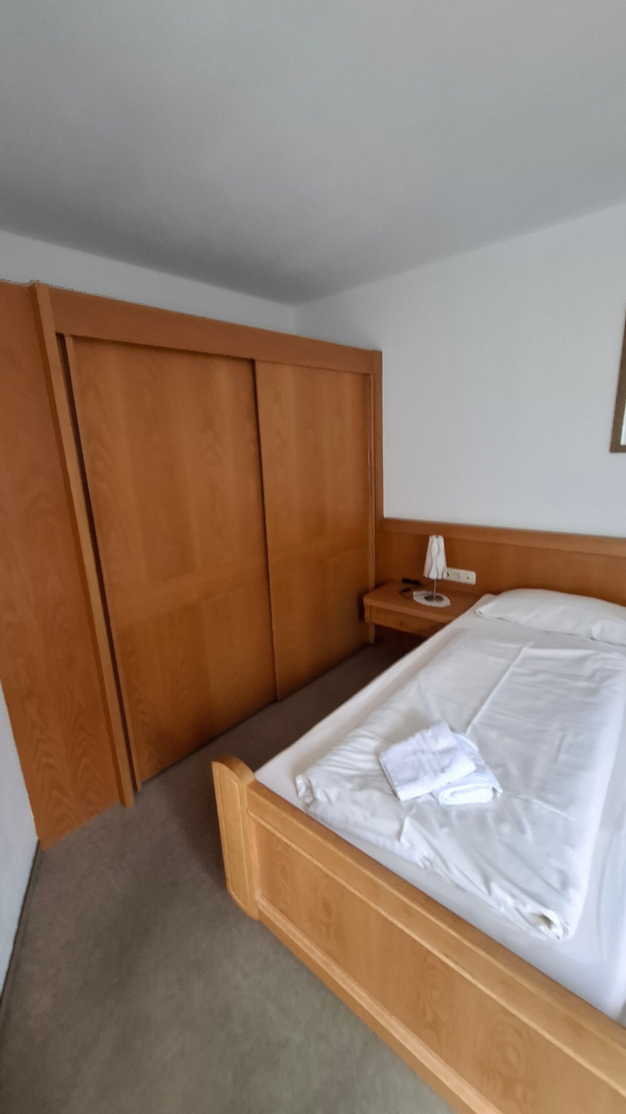 A hotel room with a wooden bed, a small bedside table with a lamp, and a wardrobe.