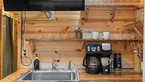 A kitchen with wooden walls, a stainless steel sink, a coffee maker, and shelves with cups and glasses. - Cedar Hollow Cabin: Cozy Black Hills Getaway (Rapid City)