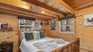 A wooden cabin bedroom with a bed, pillows, a fireplace, and framed pictures on the walls. - Cedar Hollow Cabin: Cozy Black Hills Getaway (Rapid City)