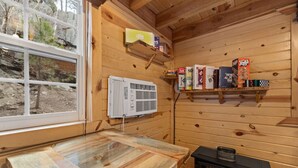 A wooden cabin interior with a wooden table, a black stove, a window, and a shelf with books and boxes. - Cedar Hollow Cabin: Cozy Black Hills Getaway (Rapid City)