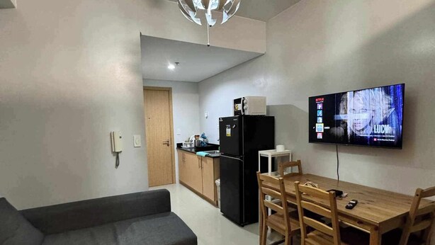 A modern living room with a sofa, dining table with chairs, a kitchen area with cabinets and appliances, and a mounted TV displaying a show. - GRASS Residences SM NORTH QC Staycation (Quezon City)