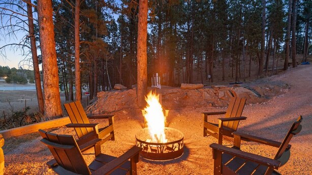 A fire pit with four wooden chairs arranged around it, set in a forested area with a dirt ground. - Bison Trail House: Rustic Cabin Retreat (Rapid City)