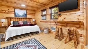 A cozy wooden cabin bedroom with a bed, a TV mounted on the wall, and a small table with stools. - Bison Trail House: Rustic Cabin Retreat (Rapid City)