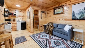 A cozy cabin interior with a wooden interior, a gray sofa, a patterned rug, and a painting of a forest on the wall. - Bison Trail House: Rustic Cabin Retreat (Rapid City)