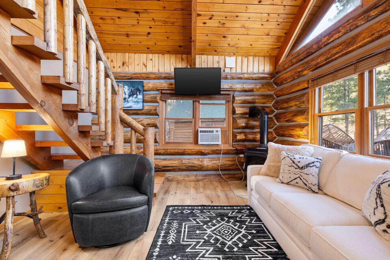 A log cabin interior with a staircase, a black leather chair, a white sofa, a black rug, and a television on the wall.
