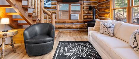 A log cabin interior with a staircase, a black leather chair, a white sofa, a black rug, and a television on the wall.