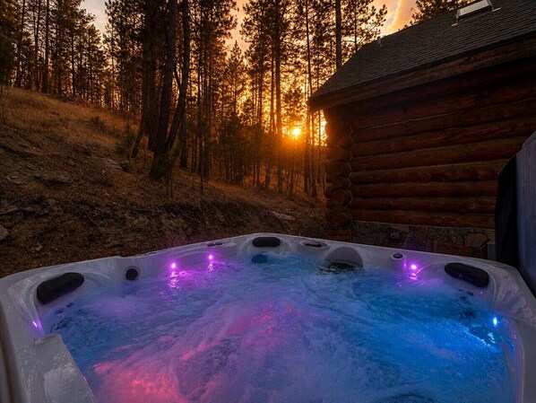 Un jacuzzi iluminado con luces LED, rodeado por un bosque y una cabaña de madera.