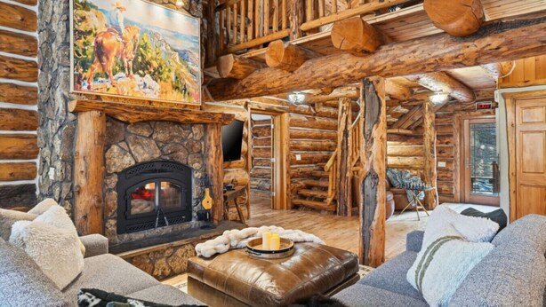 A log cabin interior with a stone fireplace, a painting above it, and a cozy seating area with a leather ottoman. - Great Lodge at Hillside: Iconic 6K Sqft Log Lodge (Rapid City)
