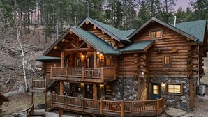 A log cabin with a stone foundation, a balcony, and a staircase leading to the upper level. - Great Lodge at Hillside: Iconic 6K Sqft Log Lodge (Rapid City)