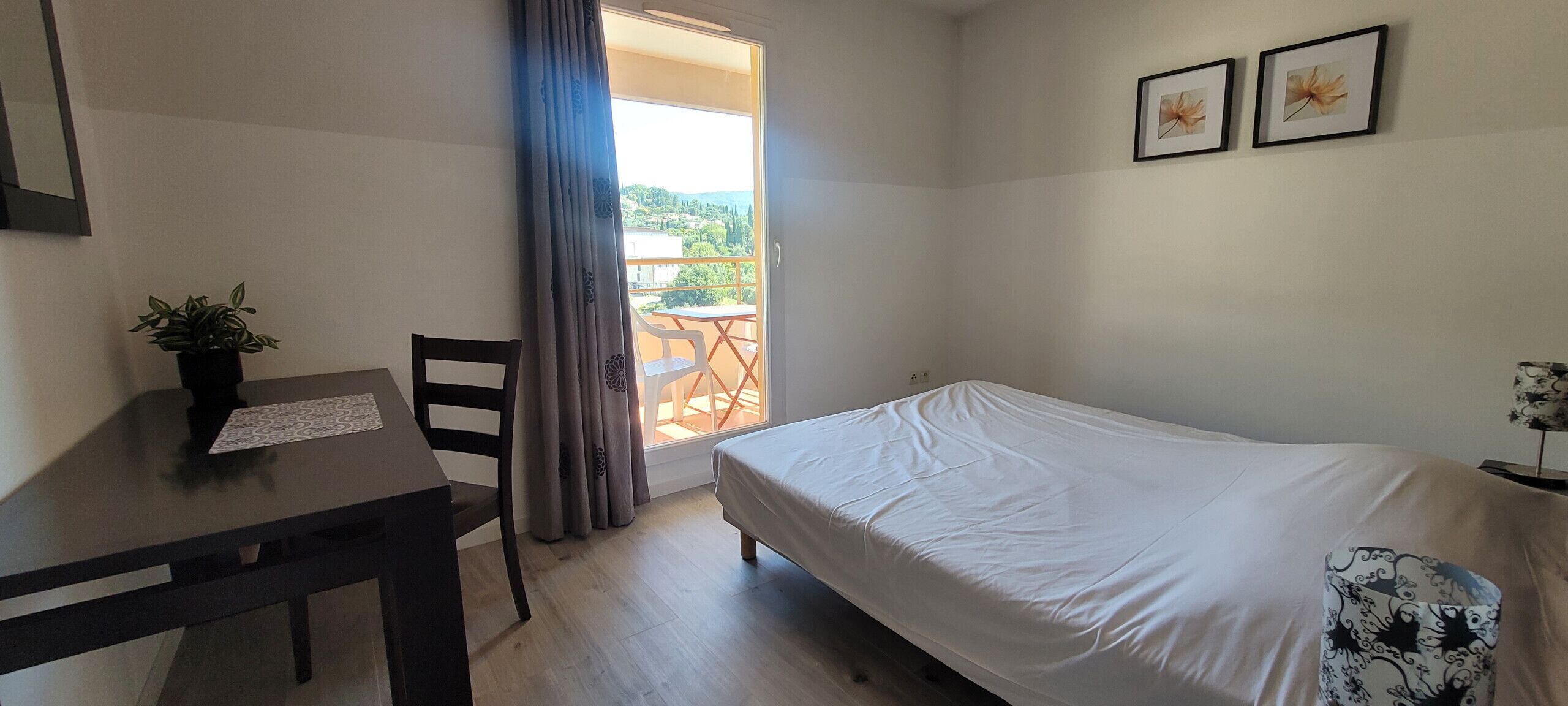 A bedroom with a bed, a desk, a chair, a potted plant, and a balcony with a view.