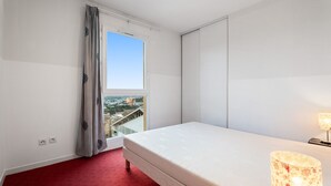 Hotellihuone, jossa on sänky, ikkuna, josta avautuu näkymä kaupungin ylle, punainen matto ja lamppu.